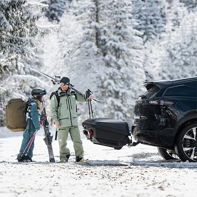 Familie med ski ved Thule Arcos XL bagageboks 450L på anhængertrækket af en sort bil i sne