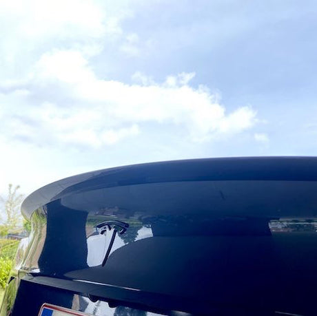 Blank sort ABS-spoiler på bagklappen af en sort Tesla Model 3 med blå himmel og skyer spejlet i lakken