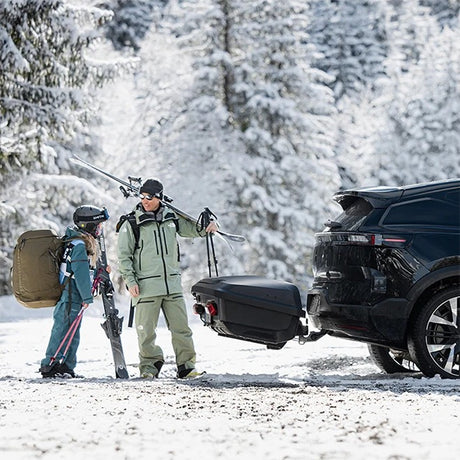 Familie med ski ved Thule Arcos XL bagageboks 450L på anhængertrækket af en sort bil i sne